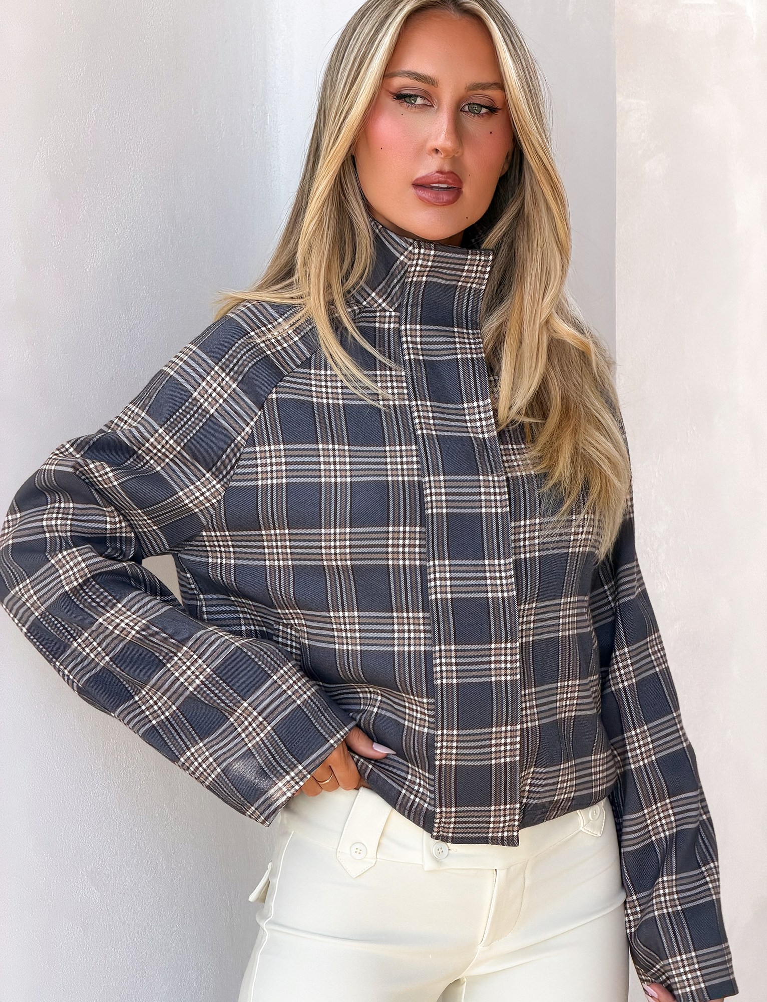 A woman with long blonde hair stands against a light wall, wearing the TIGERMIST LENNEX JACKET in grey check, which features an oversized fit and a mock neck collar, paired with white pants.