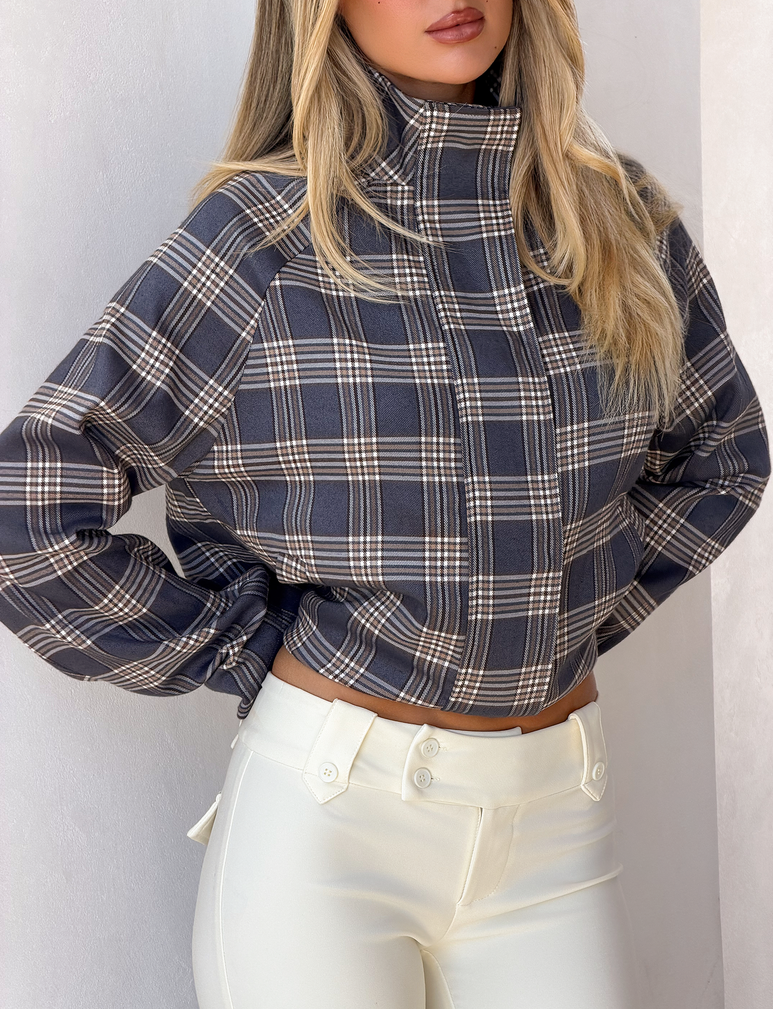 A woman stands against a light wall in the TIGERMIST LENNEX JACKET - GREY CHECK, featuring an oversized fit, mock neck collar, and long sleeves, paired with high-waisted white pants. Her loose blonde hair frames her partially visible face.
