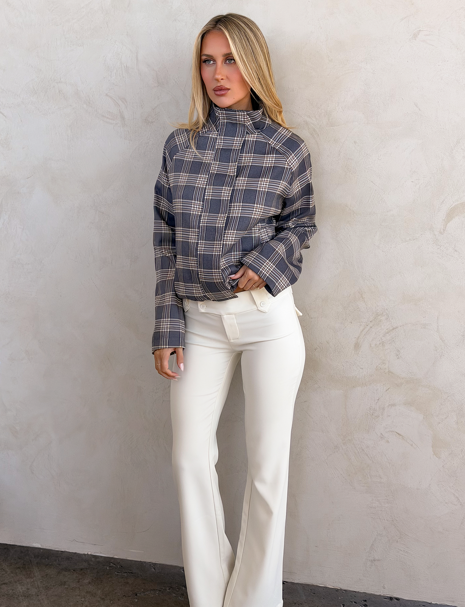 A woman stands in front of a wall, wearing the TIGERMIST LENNEX JACKET in grey check with a mock neck collar.