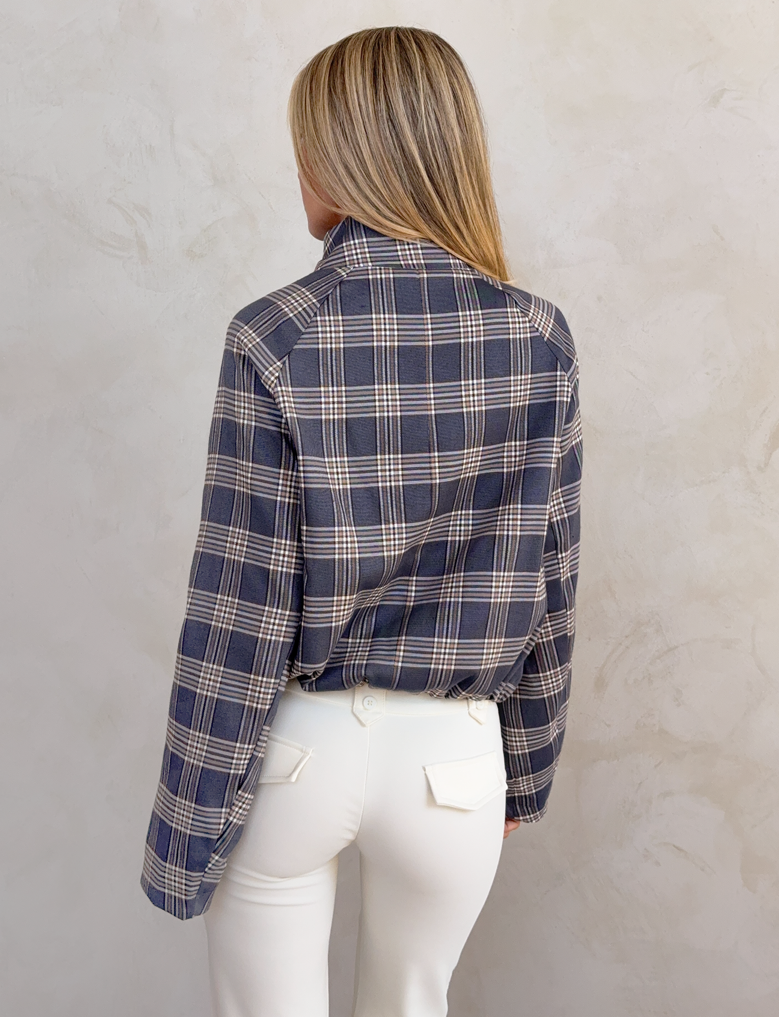 Someone with straight blonde hair is seen from behind, wearing the TIGERMIST LENNEX JACKET in grey check with a mock neck collar and white pants, standing against a light textured wall.