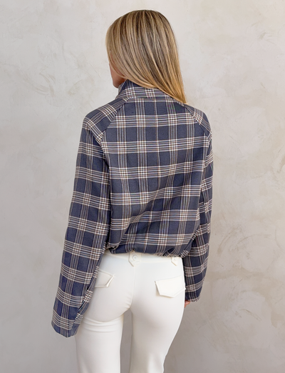 Someone with straight blonde hair is seen from behind, wearing the TIGERMIST LENNEX JACKET in grey check with a mock neck collar and white pants, standing against a light textured wall.