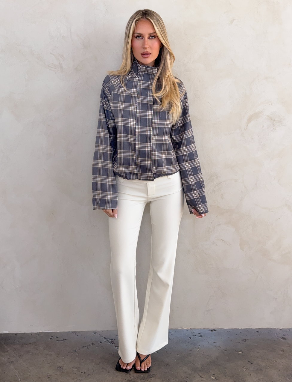 A woman with long blonde hair stands against a light textured wall, wearing the TIGERMIST LENNEX JACKET in grey check, paired with white flared pants and black sandals. Her expression is neutral and her hands rest at her sides.