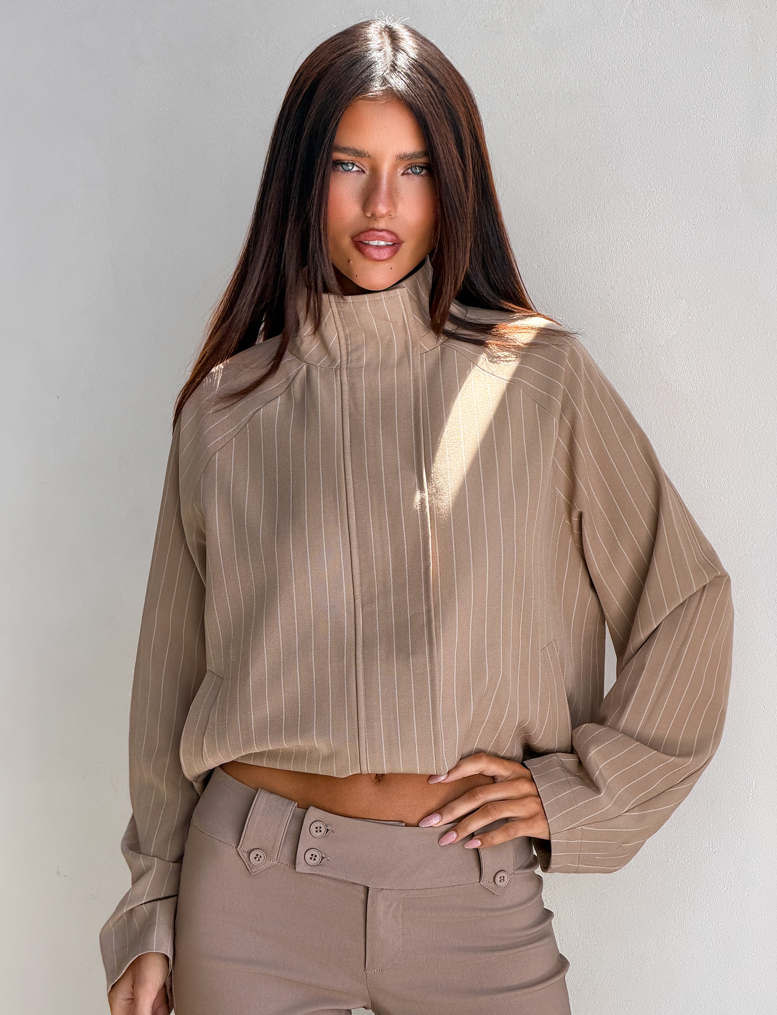 A woman with long brown hair poses against a light wall, wearing the TIGERMIST LENNEX JACKET in tan pinstripe with high-waisted beige pants and one hand on her hip.
