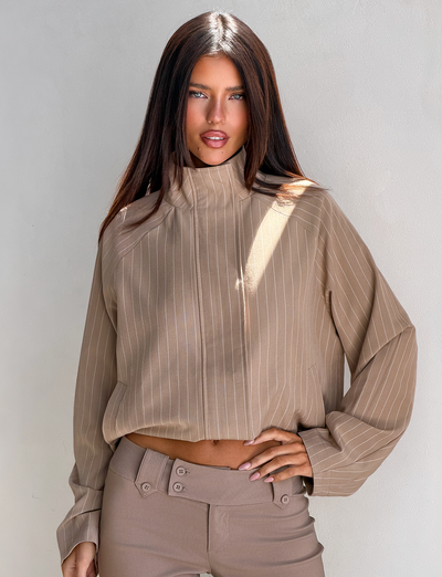 A woman with long brown hair poses against a light wall, wearing the TIGERMIST LENNEX JACKET in tan pinstripe with high-waisted beige pants and one hand on her hip.