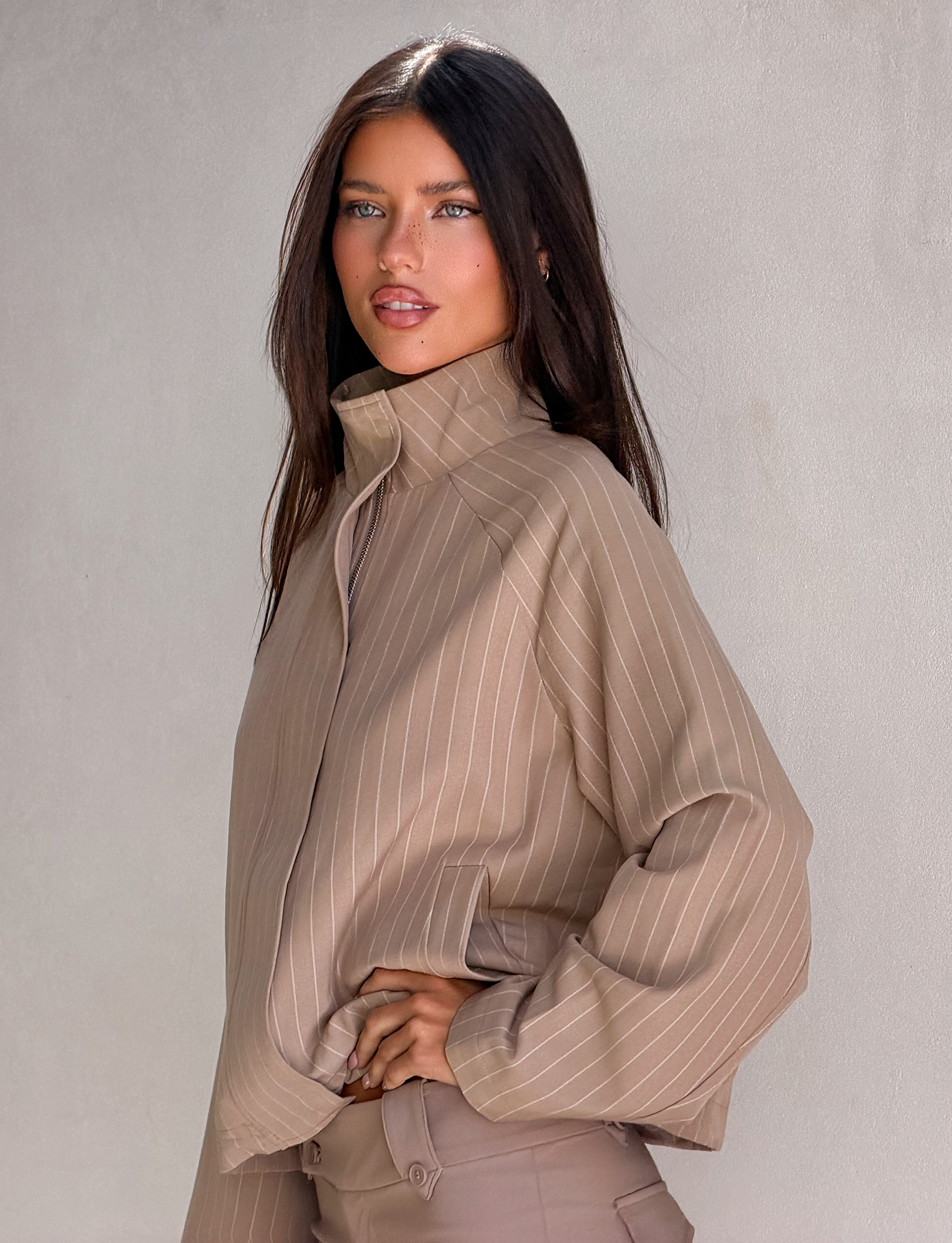 A woman with long dark hair poses confidently against a light gray wall, wearing the TIGERMIST LENNEX JACKET in tan pinstripe with her hand on her hip.
