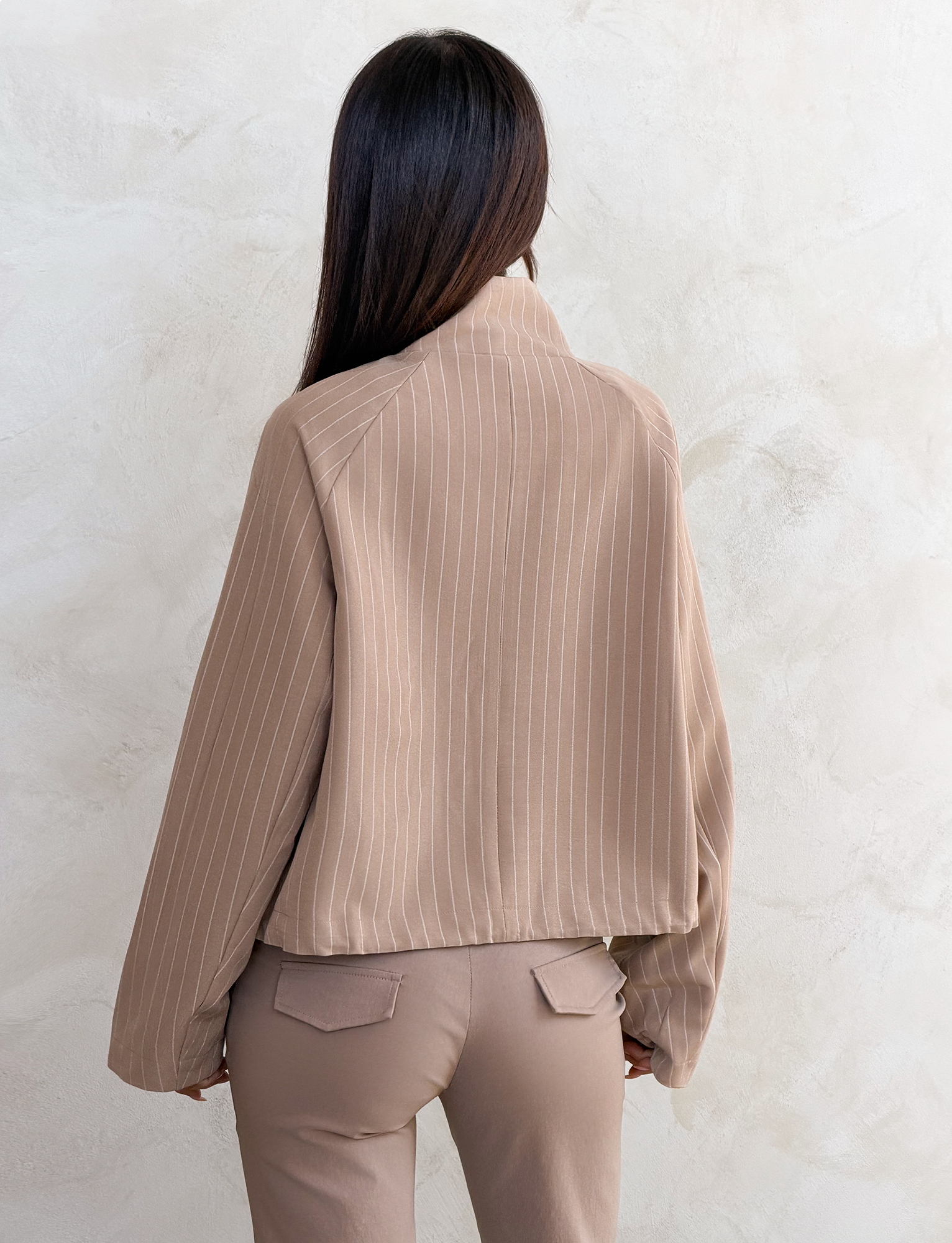 A woman with straight dark hair, seen from behind, wears the TIGERMIST LENNEX JACKET in tan pinstripe with matching pants, creating a chic look as she stands against a textured light-colored wall.