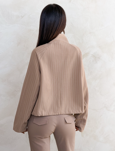 A woman with straight dark hair, seen from behind, wears the TIGERMIST LENNEX JACKET in tan pinstripe with matching pants, creating a chic look as she stands against a textured light-colored wall.