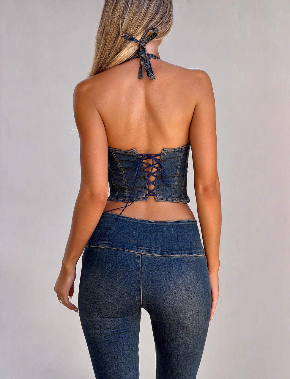 A woman with long, light brown hair is shown from the back wearing the TIGERMIST RIONA TOP - MIDWASH, a dark blue denim corset halter top, paired with high-waisted jeans against a plain light-colored background.