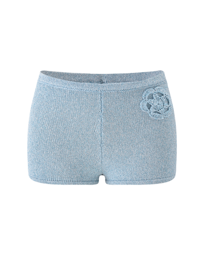 The SUNIA SHORT - BLUE by TIGERMIST are light blue ultra-mini fitted shorts with a decorative crocheted flower appliqué on the left hip, shown against a plain white background.
