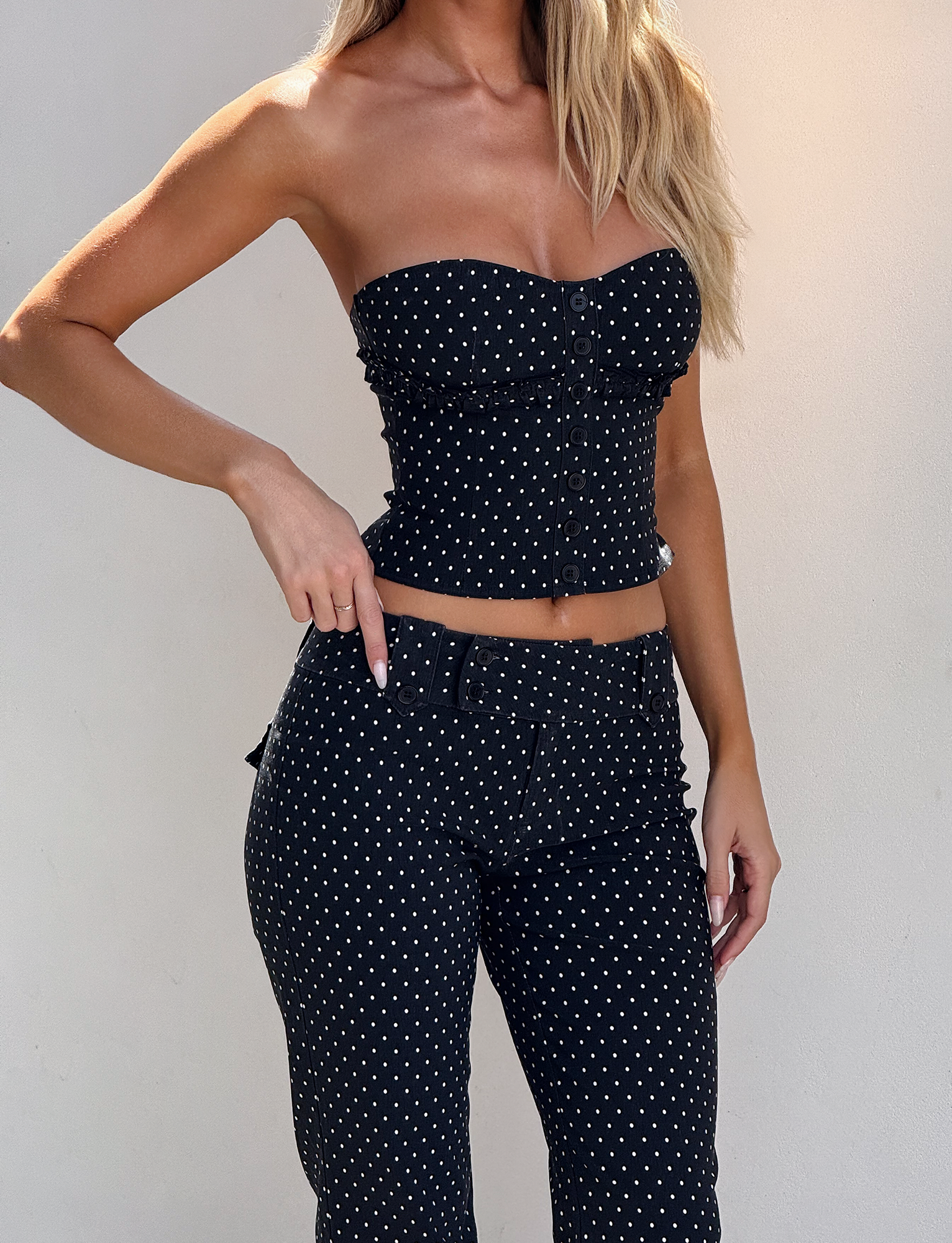 A woman with blonde hair down wears the TIGERMIST KITTIE STRAPLESS CORSET - BLACK DOT and matching high-waisted pants, standing against a light background with one hand on her belt.