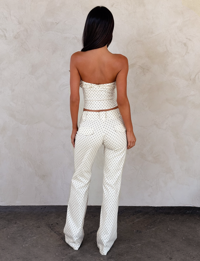A woman with long, dark hair stands facing a textured beige wall, wearing the TIGERMIST KITTIE PANT - CREAM DOT and a matching strapless polka dot top, both made from stretch suiting fabric, as she is photographed from behind.