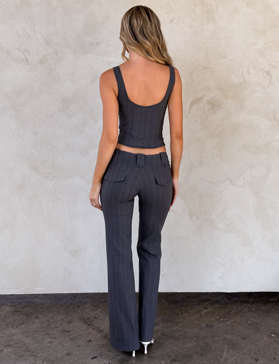 A woman with wavy blonde hair faces away, wearing TIGERMIST's KITTIE PANT - GREY STRIPE and a matching sleeveless pinstripe top in stretch suiting fabric, standing on a dark floor against a light textured wall.