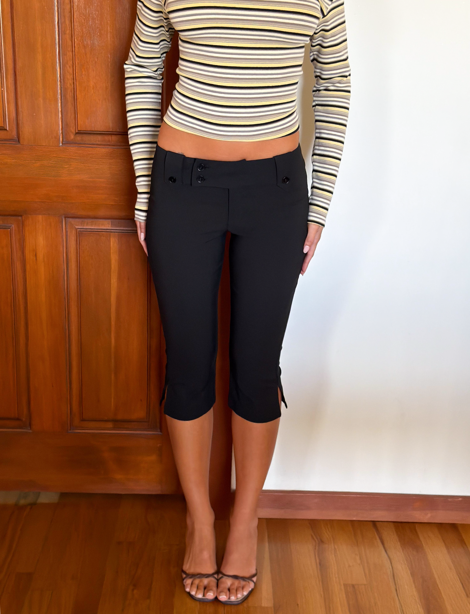 A person stands indoors in front of a wooden door and hardwood floor, wearing the TIGERMIST KITTIE CAPRI - BLACK with side slits and a crossover waistband, paired with a long-sleeve striped crop top. Their head and feet are not visible.