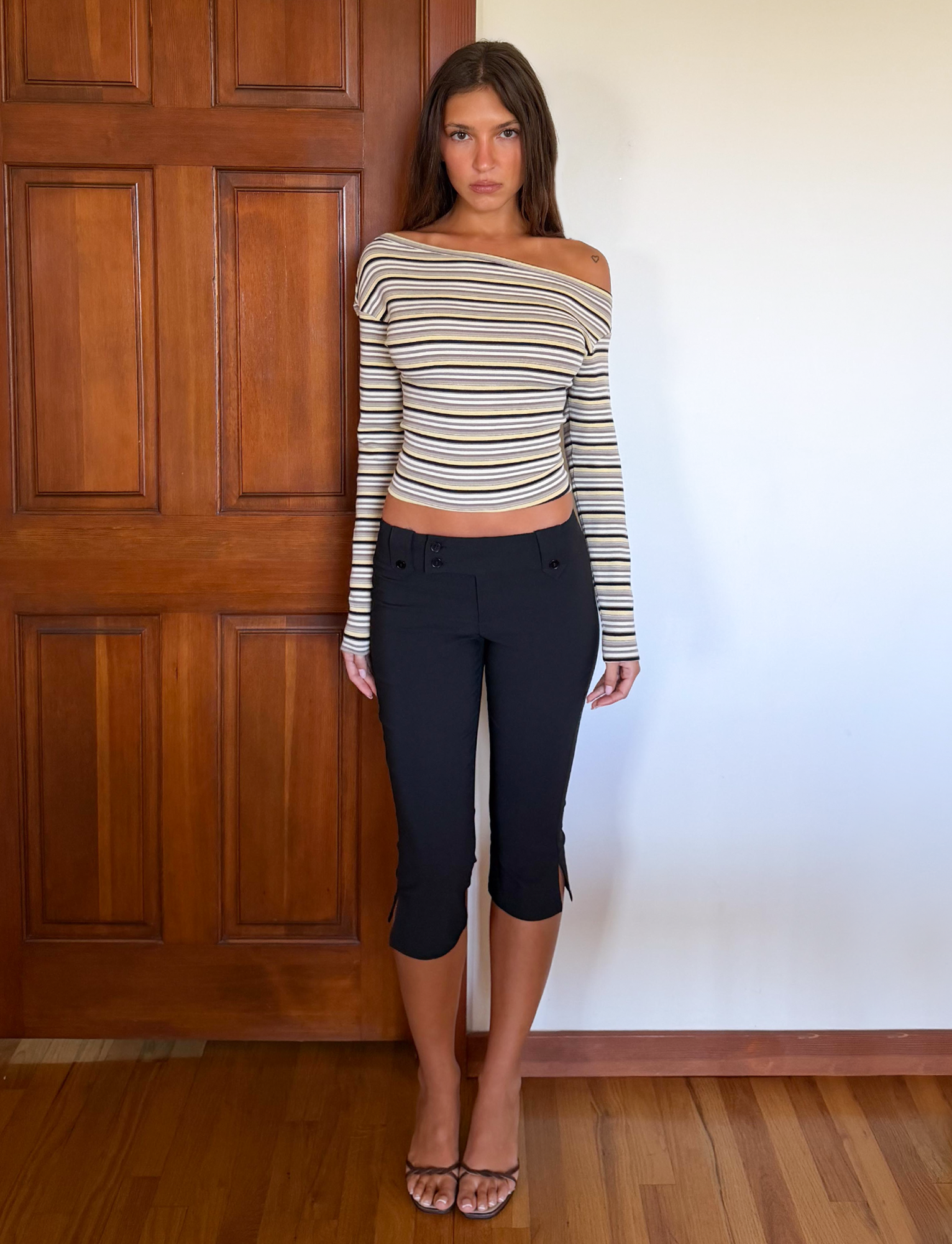 A woman stands indoors in front of a wooden door, wearing a striped off-the-shoulder top, TIGERMIST KITTIE CAPRI - BLACK pants with a crossover waistband, and clear-strap heels on a hardwood floor.