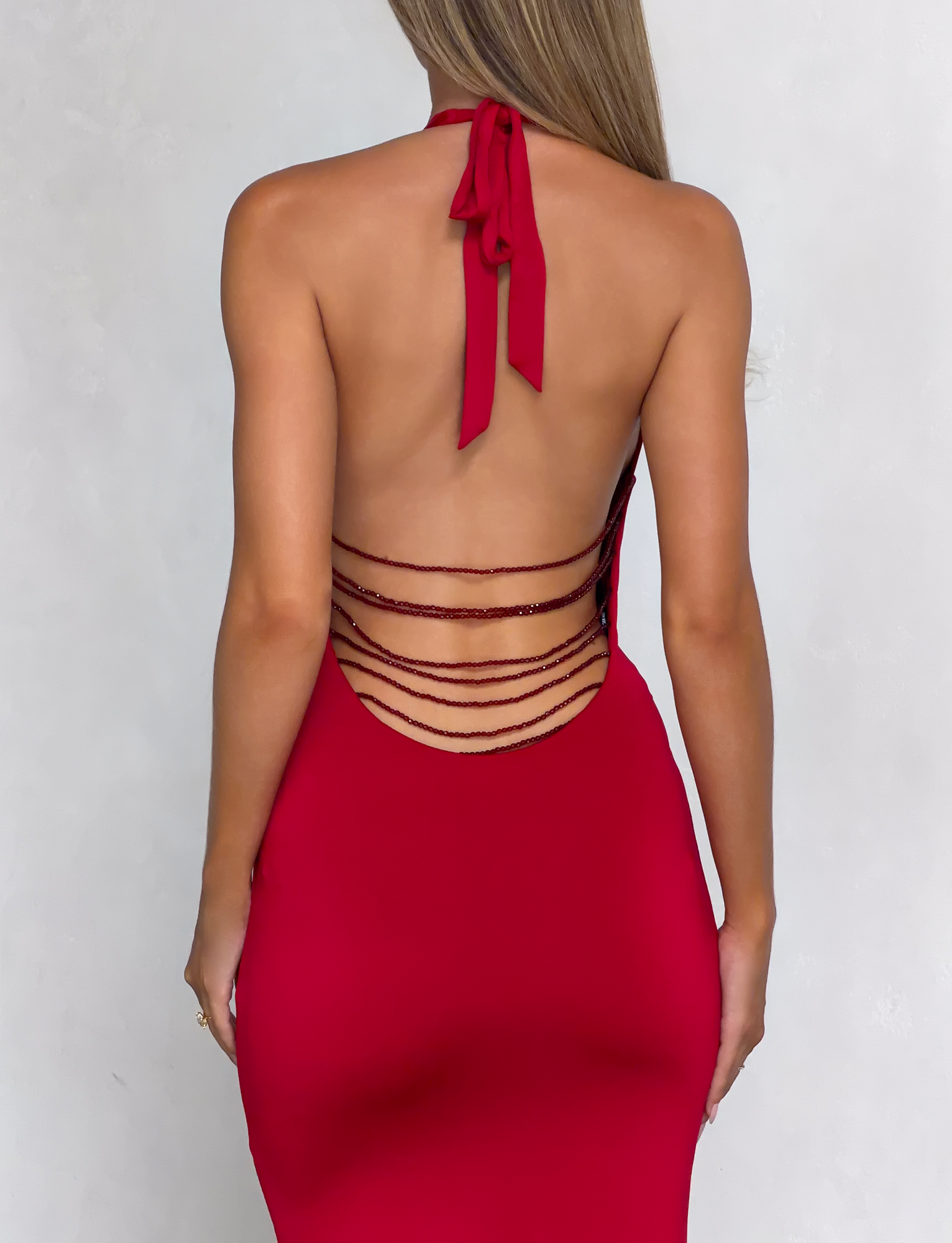 A woman wearing the TIGERMIST SOLYNE MAXI DRESS in red, featuring a backless halter neck design with multiple thin horizontal straps across the lower back, stands against a light-colored background.