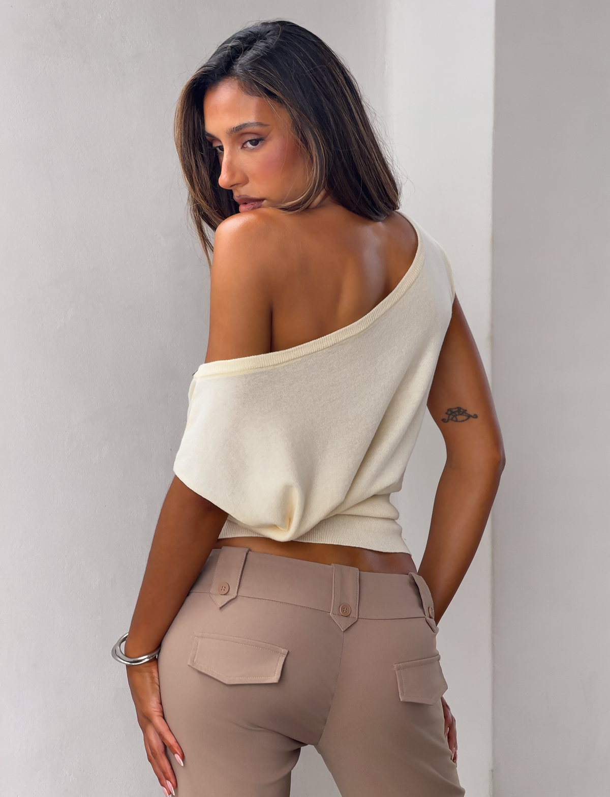 A woman with loose hair and a bracelet poses with her back to the camera, wearing the TIGERMIST TYLA SLEEVELESS KNIT TOP in cream and beige pants, standing against a light-colored wall.