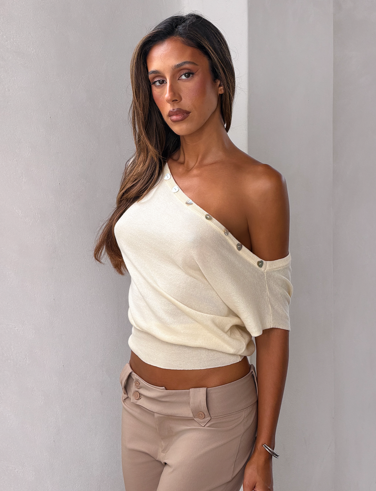 A woman with long brown hair wears the TIGERMIST TYLA SLEEVELESS KNIT TOP in cream, featuring heart shell buttons, paired with beige pants. She stands against a light wall and looks confidently at the camera.