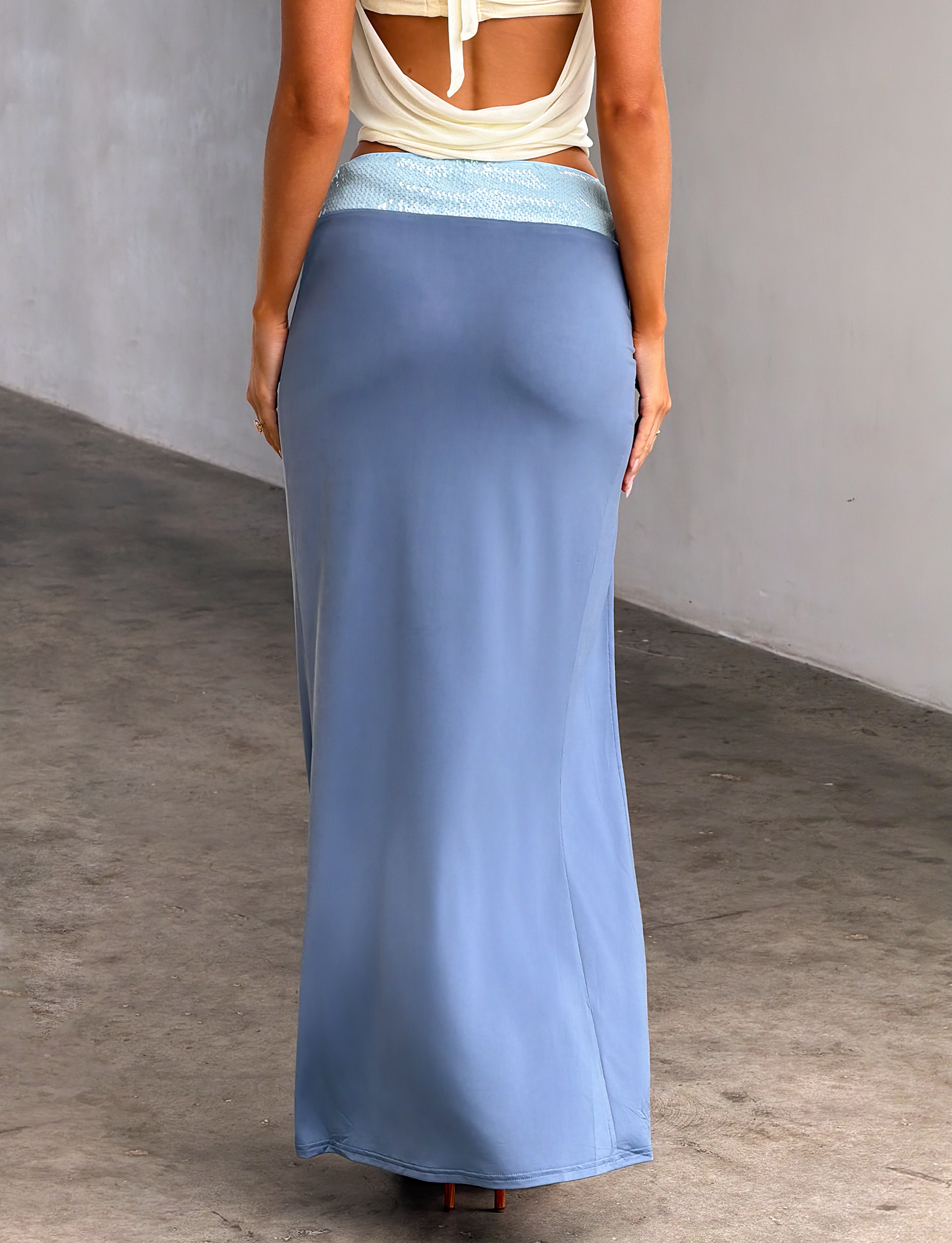 A woman faces away from the camera, wearing a cream sleeveless top and the TIGERMIST THARA MAXI SKIRT in slate blue. She stands indoors against a plain light wall and concrete floor.