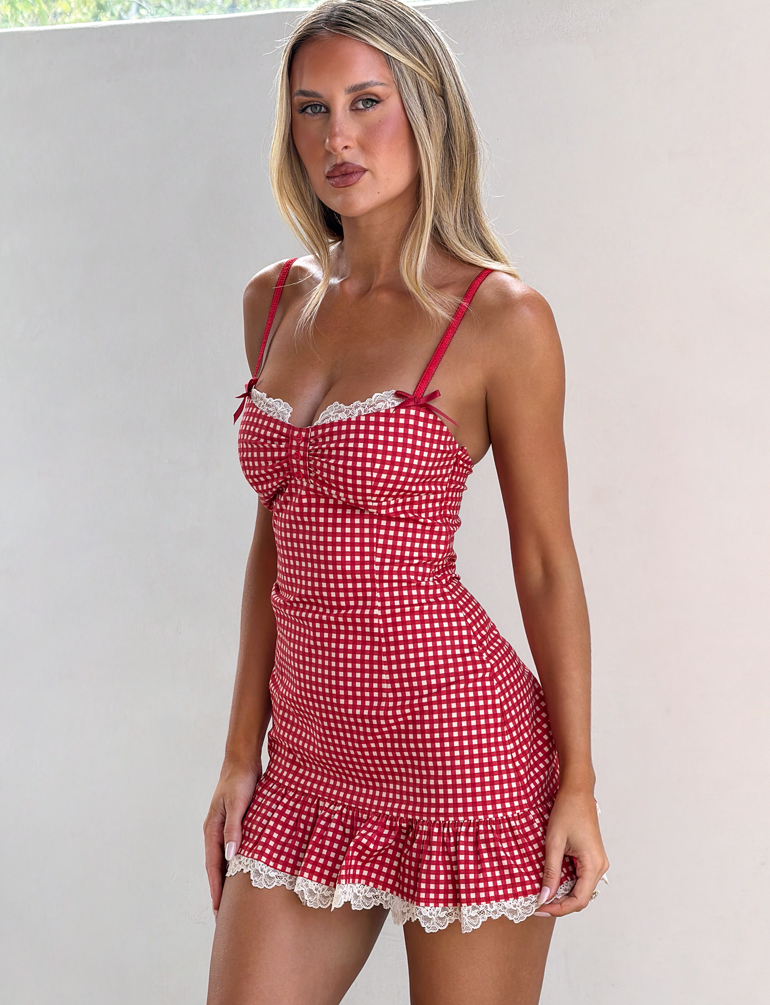 A woman wears the TIGERMIST EMMALINE MINI DRESS in red gingham with lace trim, standing against a plain light-colored wall and looking at the camera with a neutral expression.
