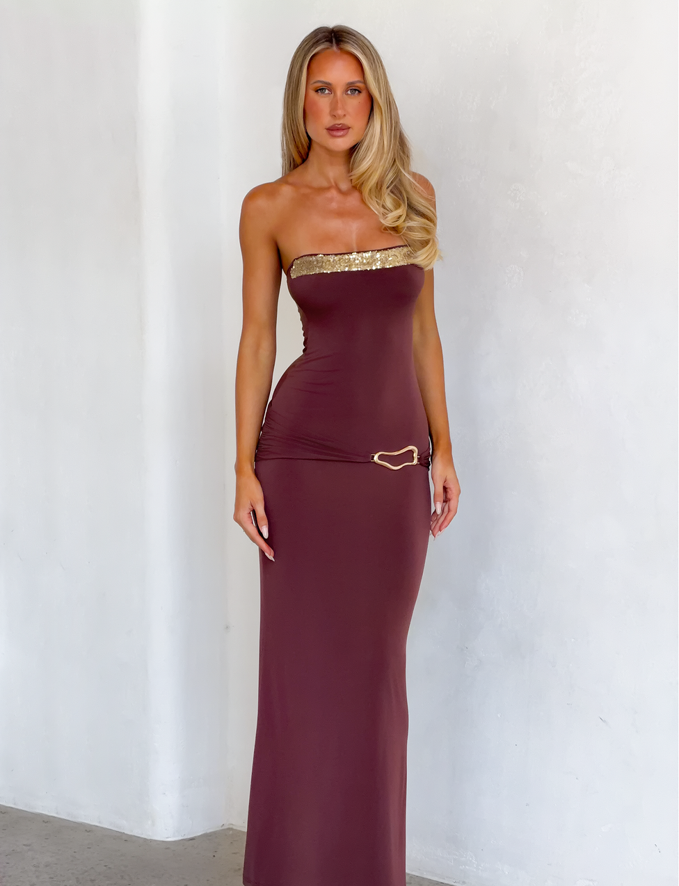 A woman with long blonde hair models the TIGERMIST EOS SEQUIN TUBE MAXI DRESS in Cinnamon Chocolate— a form-fitting, strapless sequin gown with gold trim and heart detail, standing against a plain white wall.