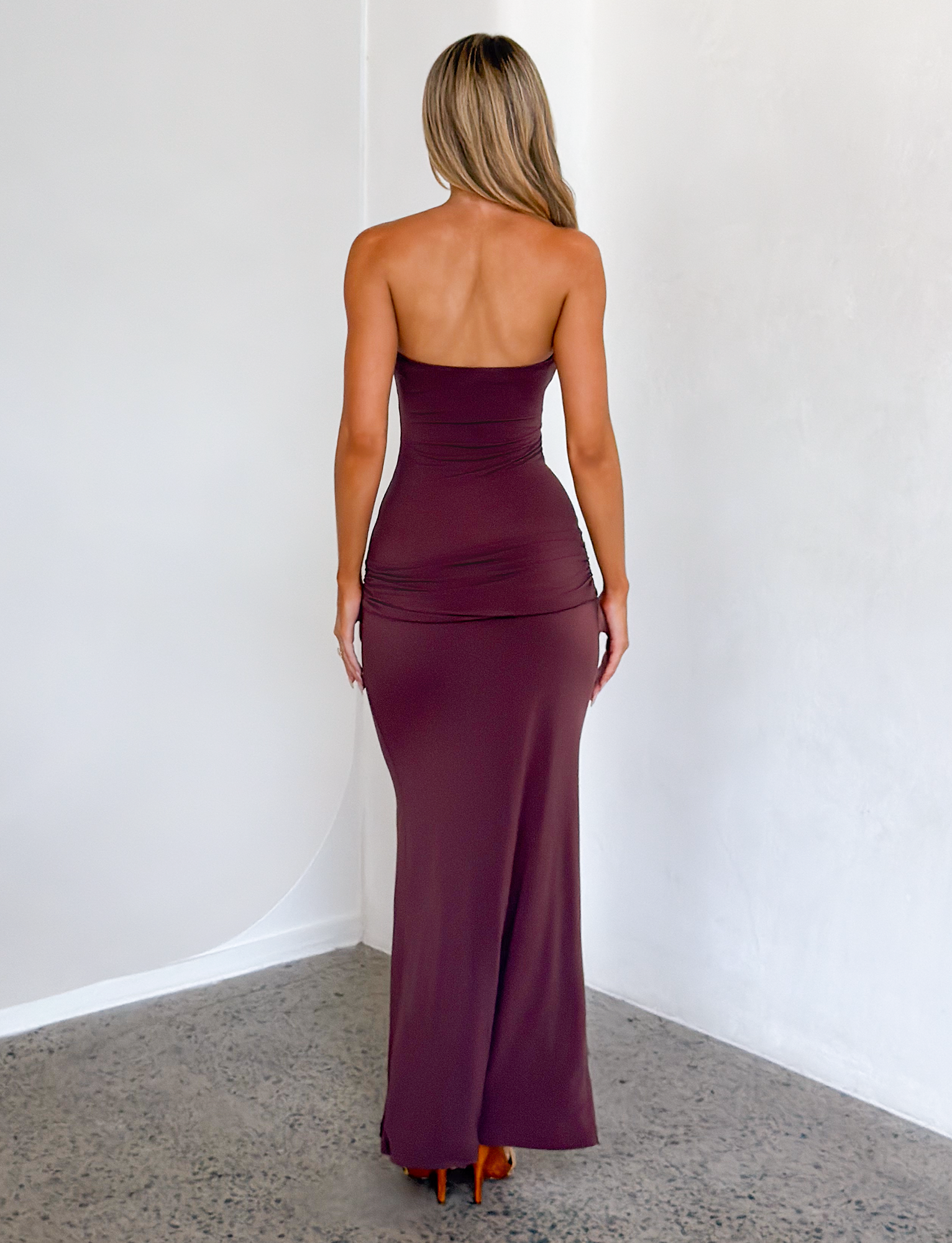 A woman with long blonde hair stands indoors on a concrete floor, facing away from the camera, wearing the TIGERMIST EOS SEQUIN TUBE MAXI DRESS in Cinnamon Chocolate. A white wall serves as the background.