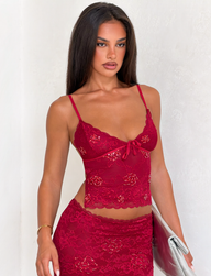 A woman with long brown hair wears the TIGERMIST KIRRA BEADED SEQUIN LACE TOP in red and a matching skirt, holding a silver clutch. She accessorizes with a thin gold bracelet and stands confidently against a light-colored wall.