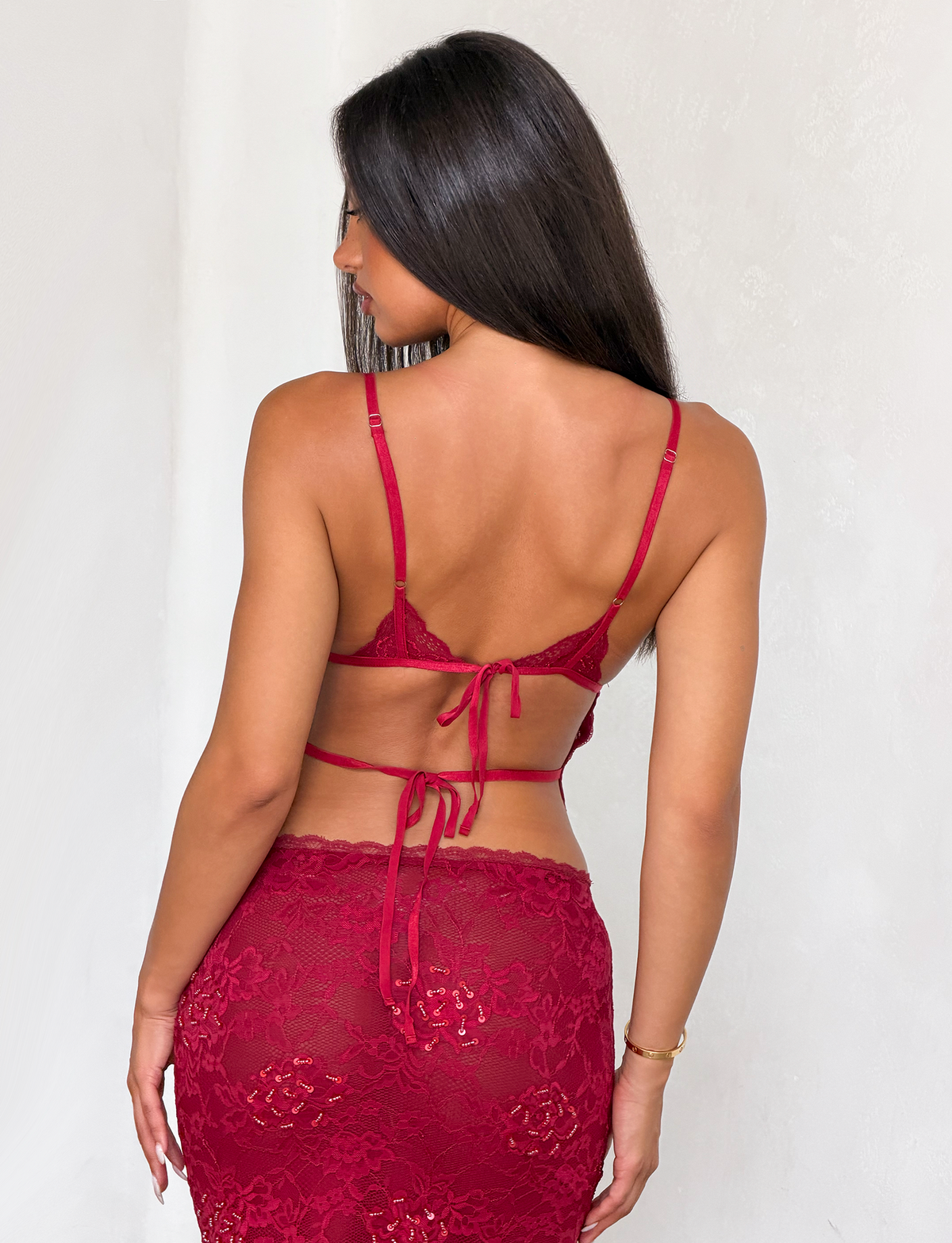A woman with long dark hair poses with her back to the camera, wearing the TIGERMIST KIRRA Beaded Sequin Lace Top in red. The top features thin straps, tie-back details, and pairs with a matching high-waisted lace skirt.