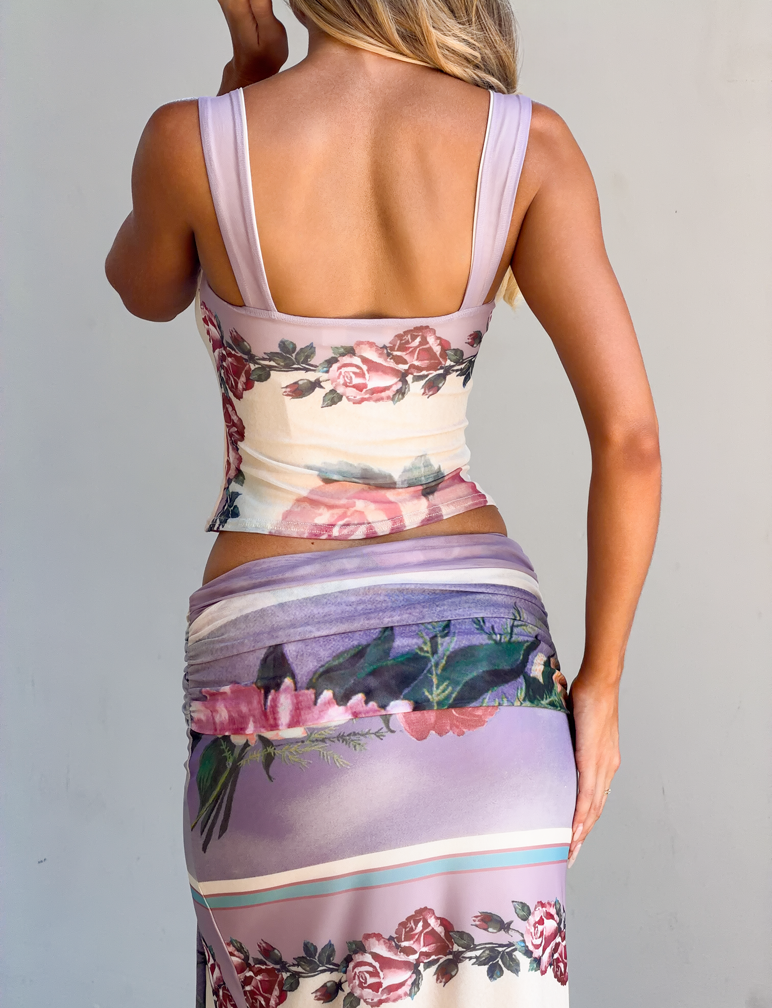 A woman faces away, wearing the TIGERMIST RAFFIE PRINTED CORSET in Postcard Floral with matching skirt in purple and pink. Her right hand is on her hip, and her long blonde hair falls to one side.
