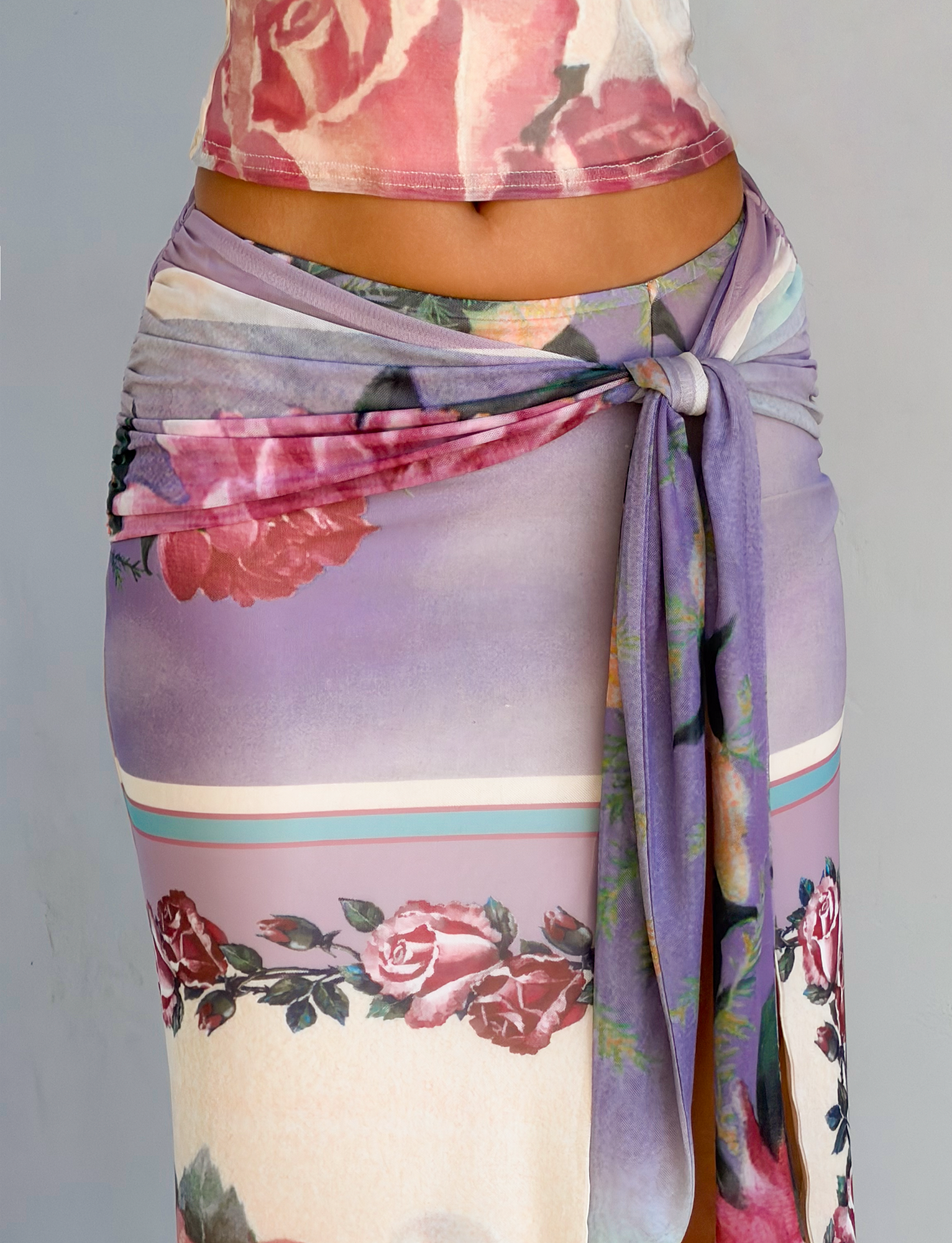 A close-up shows a person wearing the TIGERMIST RAFFIE MAXI SKIRT - POSTCARD FLORAL, a fitted purple and cream skirt with red rose prints, gathered waist tie, and high leg split, paired with a matching top against a light gray background.