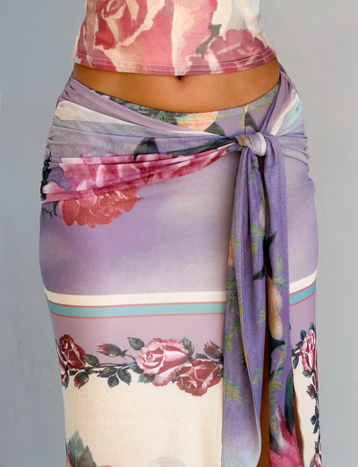 A close-up shows a person wearing the TIGERMIST RAFFIE MAXI SKIRT - POSTCARD FLORAL, a fitted purple and cream skirt with red rose prints, gathered waist tie, and high leg split, paired with a matching top against a light gray background.