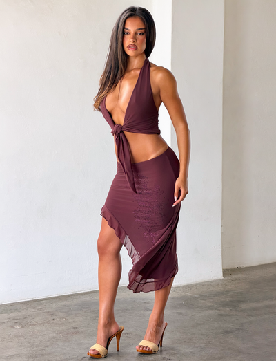 A woman with long brown hair stands indoors near a white wall, wearing TIGERMIST's GEORGIE DIAMANTE HI LO SKIRT in chocolate with a matching halter top and tan high-heeled sandals.