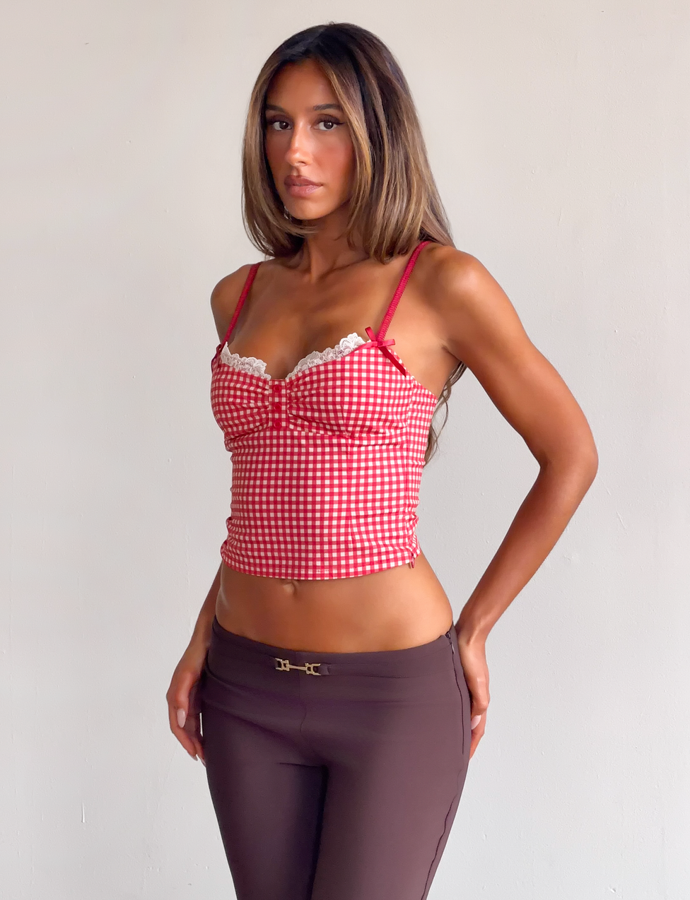 A woman wears the TIGERMIST EMMALINE BUSTIER in red gingham—featuring lace trim and adjustable straps—paired with brown pants, posing against a plain light background and looking at the camera with a neutral expression.