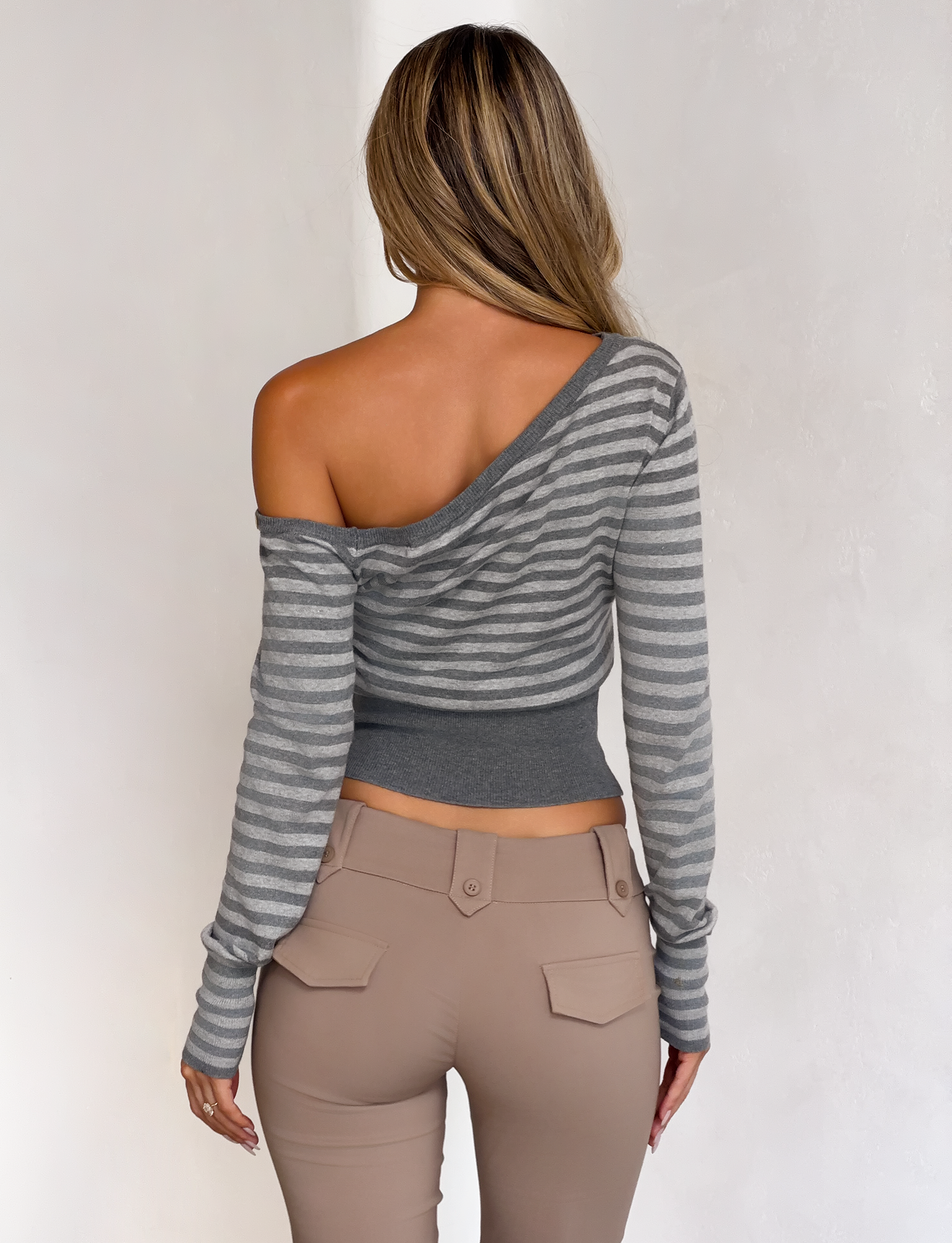 A woman with loose long hair faces away, wearing the TIGERMIST TYLA JUMPER in grey stripe with an off-the-shoulder style and high-waisted beige pants, set against a plain light wall.