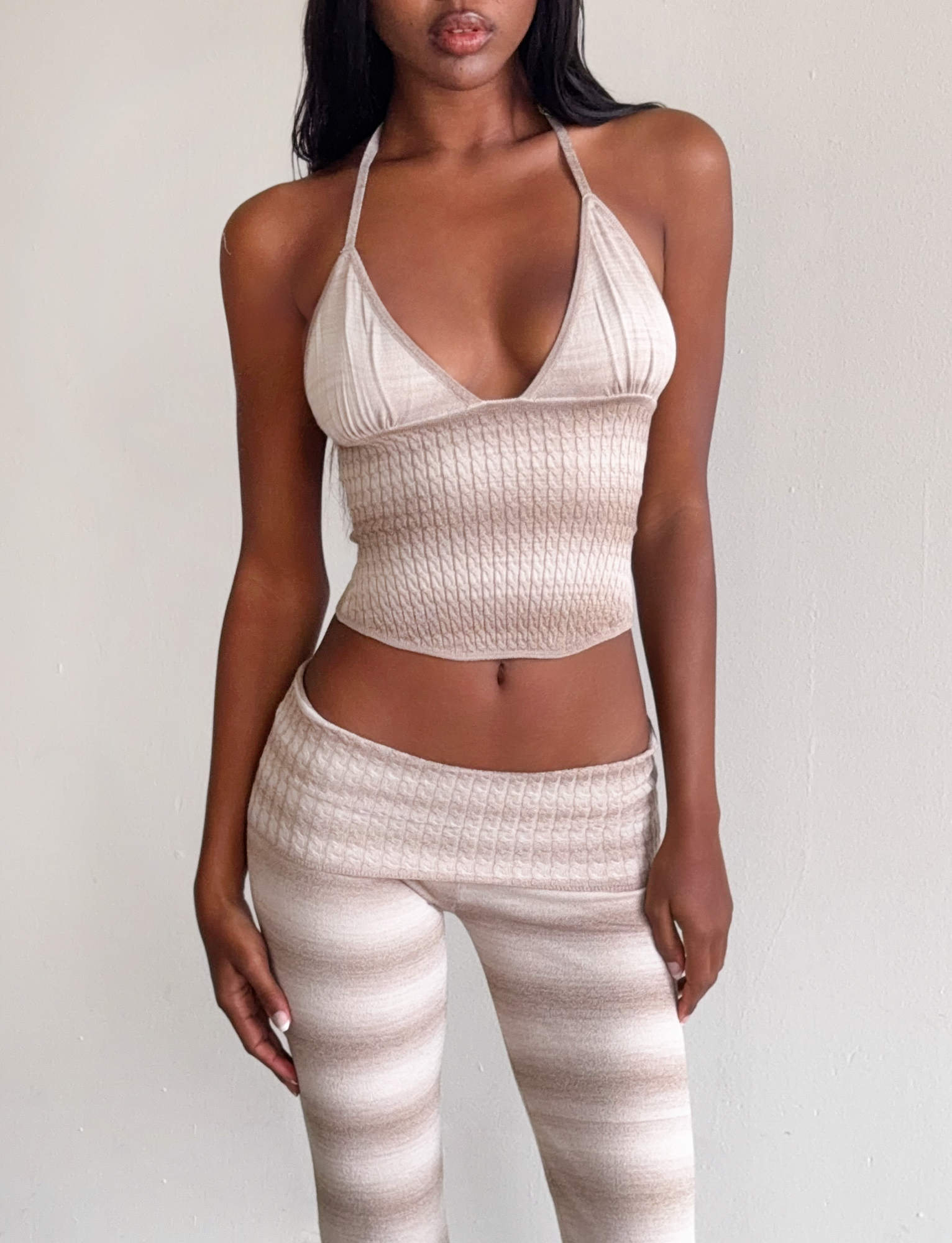 A woman poses against a plain wall wearing the TIGERMIST MARISSA KNIT CAMI in taupe ombre stripe with matching high-waisted, ribbed knit pants featuring subtle stripes. Her hands rest at her sides.