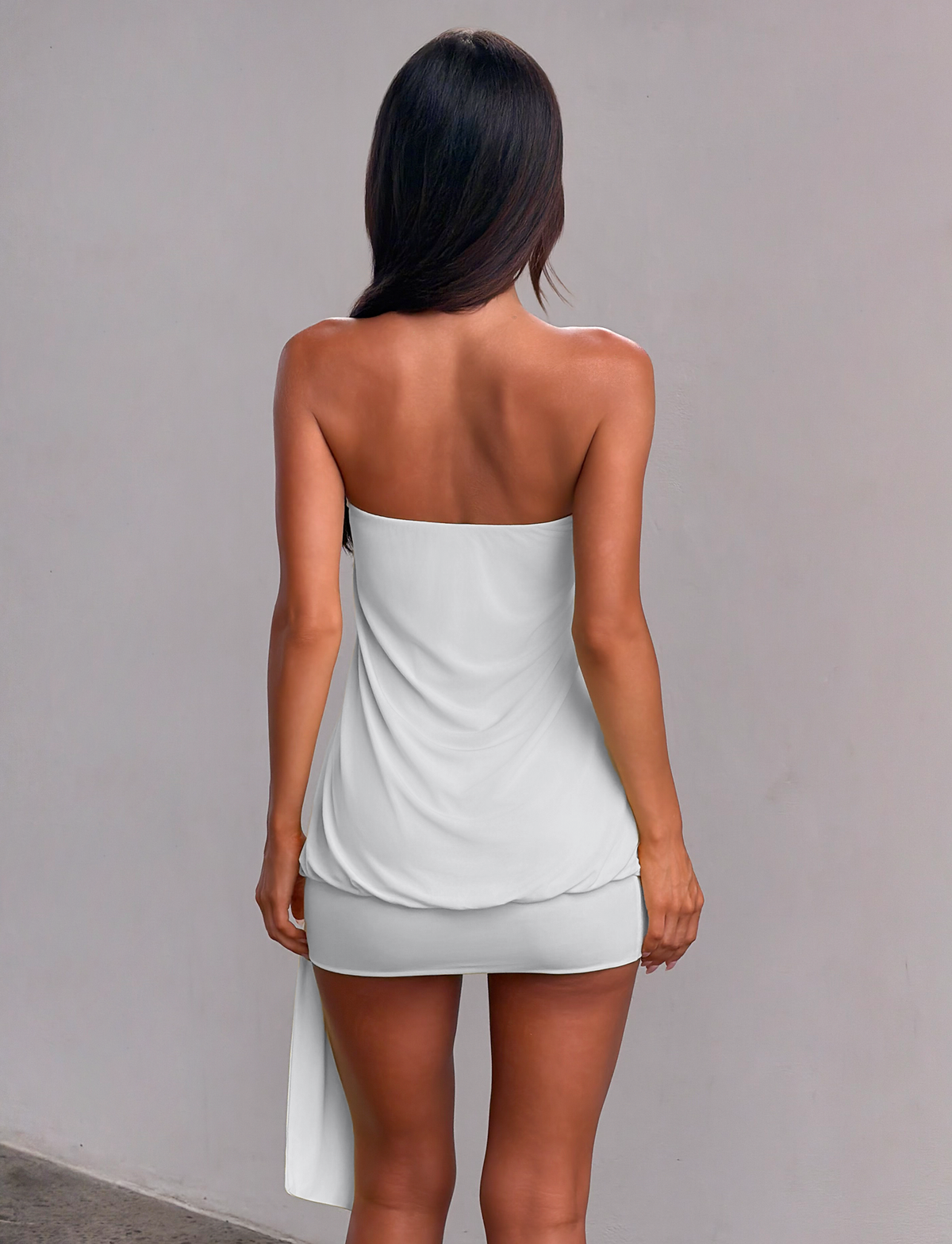 A woman with long dark hair stands facing away in the TIGERMIST RAVENNA MINI DRESS in Cool Slate, a strapless bodycon drop-waist mini, set against a plain light gray wall.