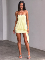 A woman with long brown hair models the TIGERMIST RAVENNA MINI DRESS in butter yellow, featuring button details and an asymmetrical drape, paired with silver high-heeled sandals against a plain gray wall.