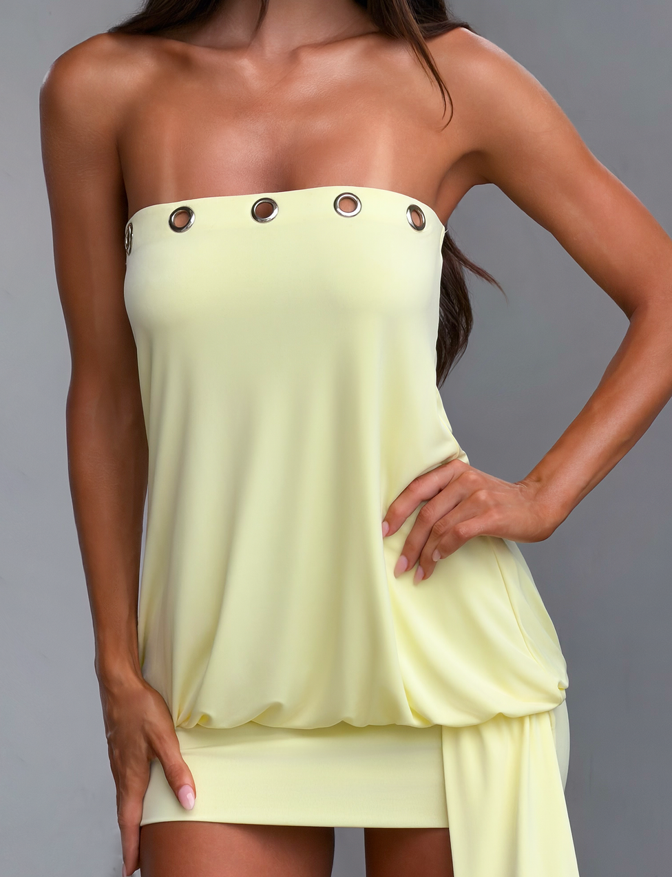 A woman models the TIGERMIST RAVENNA MINI DRESS in butter yellow—a strapless, bodycon mini with silver grommet accents and a drop waist. She poses with one hand on her hip, the other on her thigh, against a plain gray background.