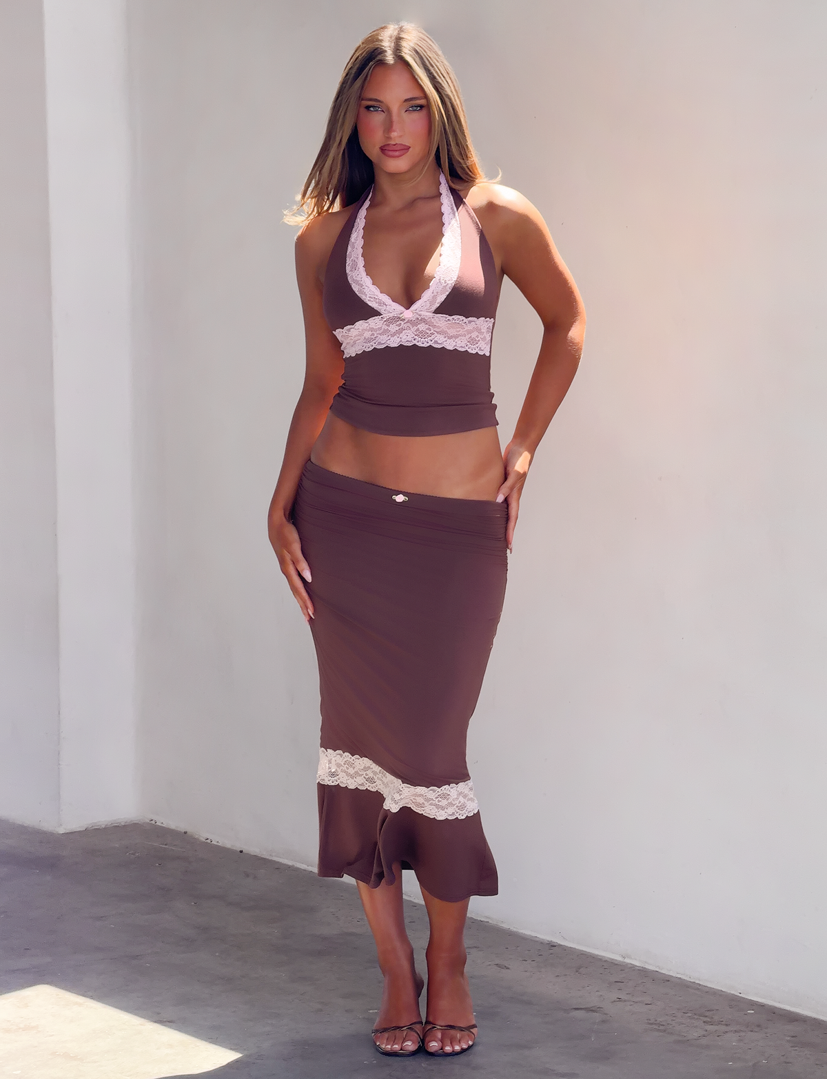 A person wearing a brown halter top with pink lace trim and a matching CALINA LACE PANEL MIDI SKIRT - CHOCOLATE with a pink lace hem.