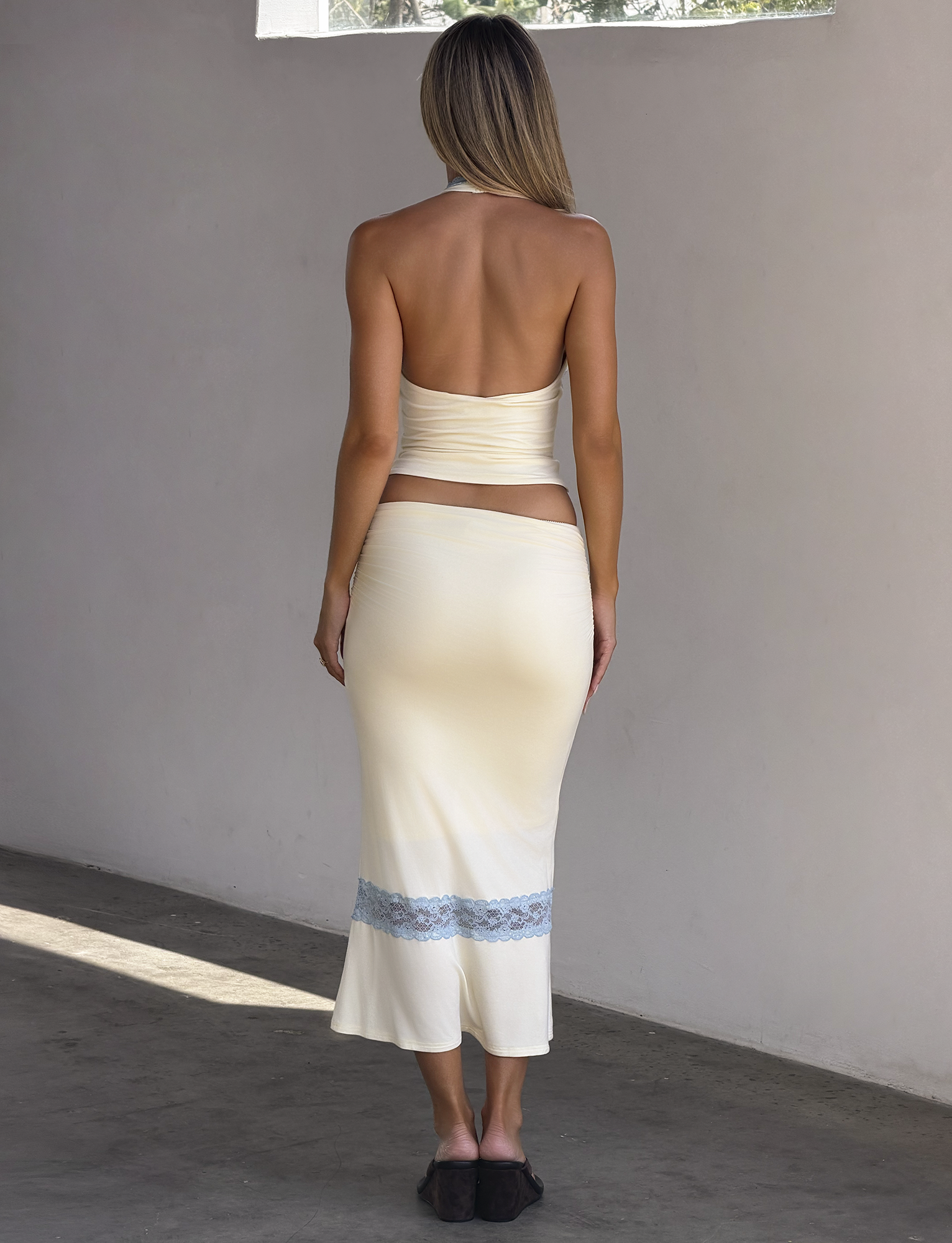 A woman stands barefoot indoors on concrete, wearing the TIGERMIST CALINA LACE PANEL MIDI SKIRT in cream with a blue lace hem and a matching backless halter top, as sunlight streams through a high window, highlighting her elegance.