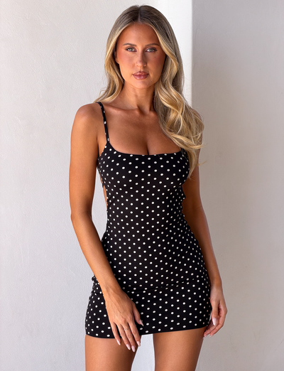 A woman with long blonde hair wears the TIGERMIST MINERVA MINI DRESS 2.0 in black polka dot, standing against a plain white wall and looking at the camera.