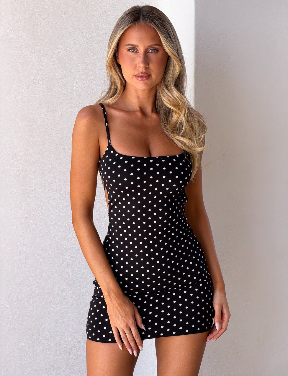 A woman with long blonde hair wears the TIGERMIST MINERVA MINI DRESS 2.0 in black polka dot, standing against a plain white wall and looking at the camera.