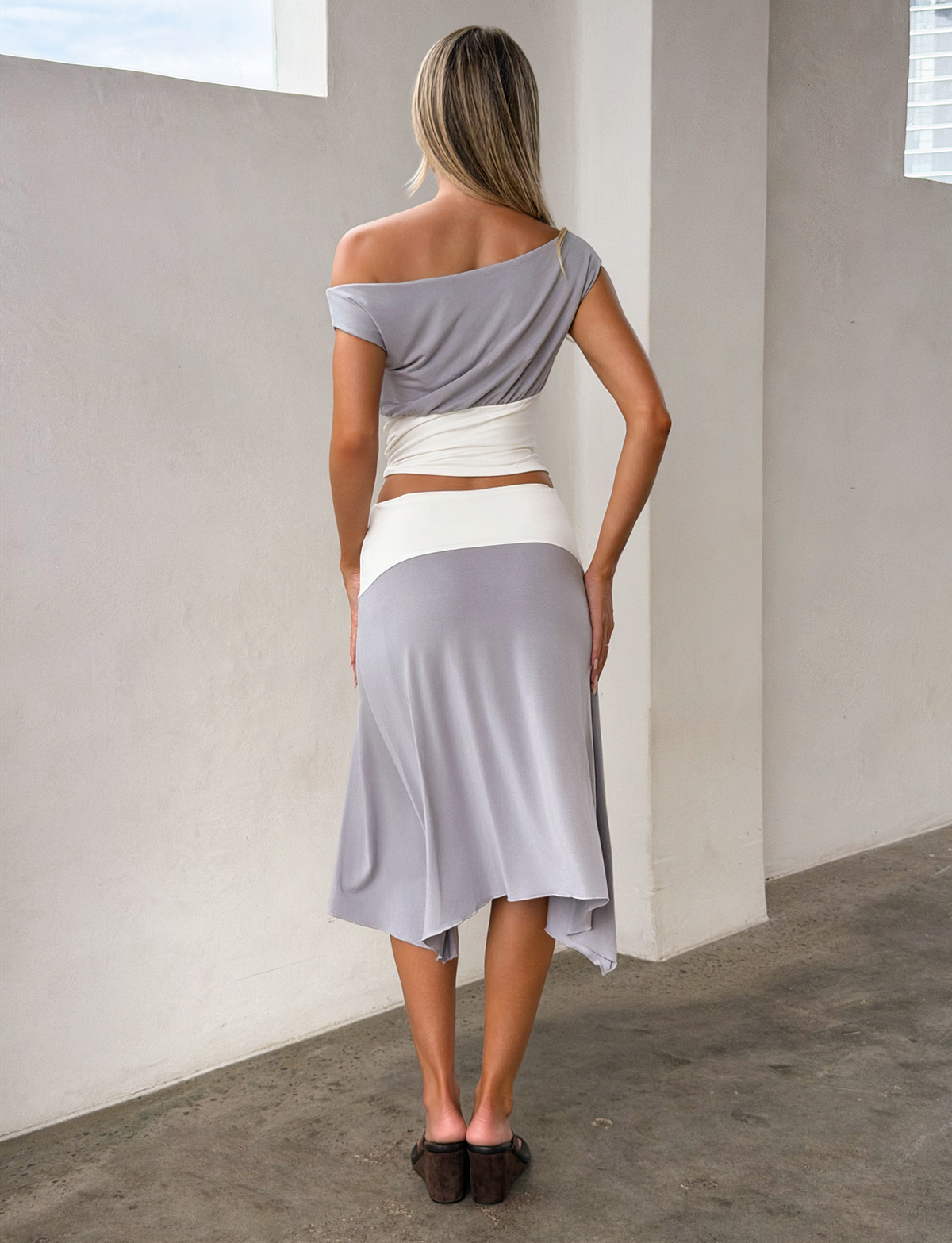 A woman with long blonde hair stands indoors, seen from behind. She wears a gray off-shoulder top, a white waist wrap, and the TIGERMIST ALYSSA HI LO SKIRT - GREY with an asymmetrical hem, paired with brown shoes.