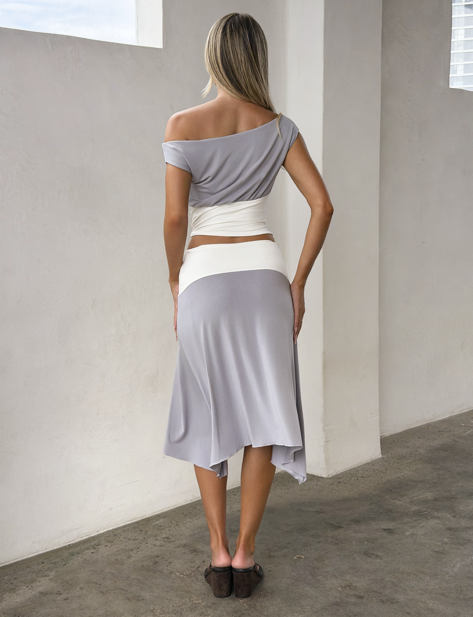 A woman with long blonde hair stands indoors, seen from behind. She wears a gray off-shoulder top, a white waist wrap, and the TIGERMIST ALYSSA HI LO SKIRT - GREY with an asymmetrical hem, paired with brown shoes.