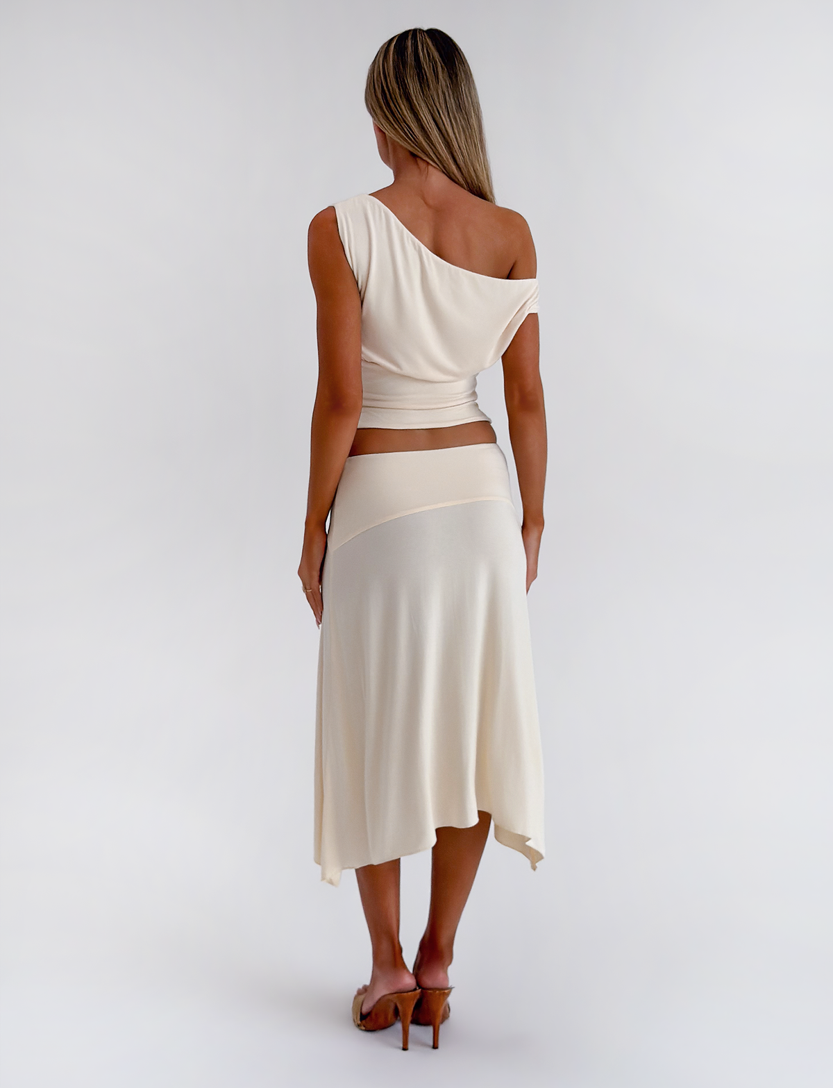 A woman faces away, wearing the TIGERMIST ALYSSA HI LO SKIRT in cream with a matching one-shoulder top and heeled sandals, set against a plain white background.
