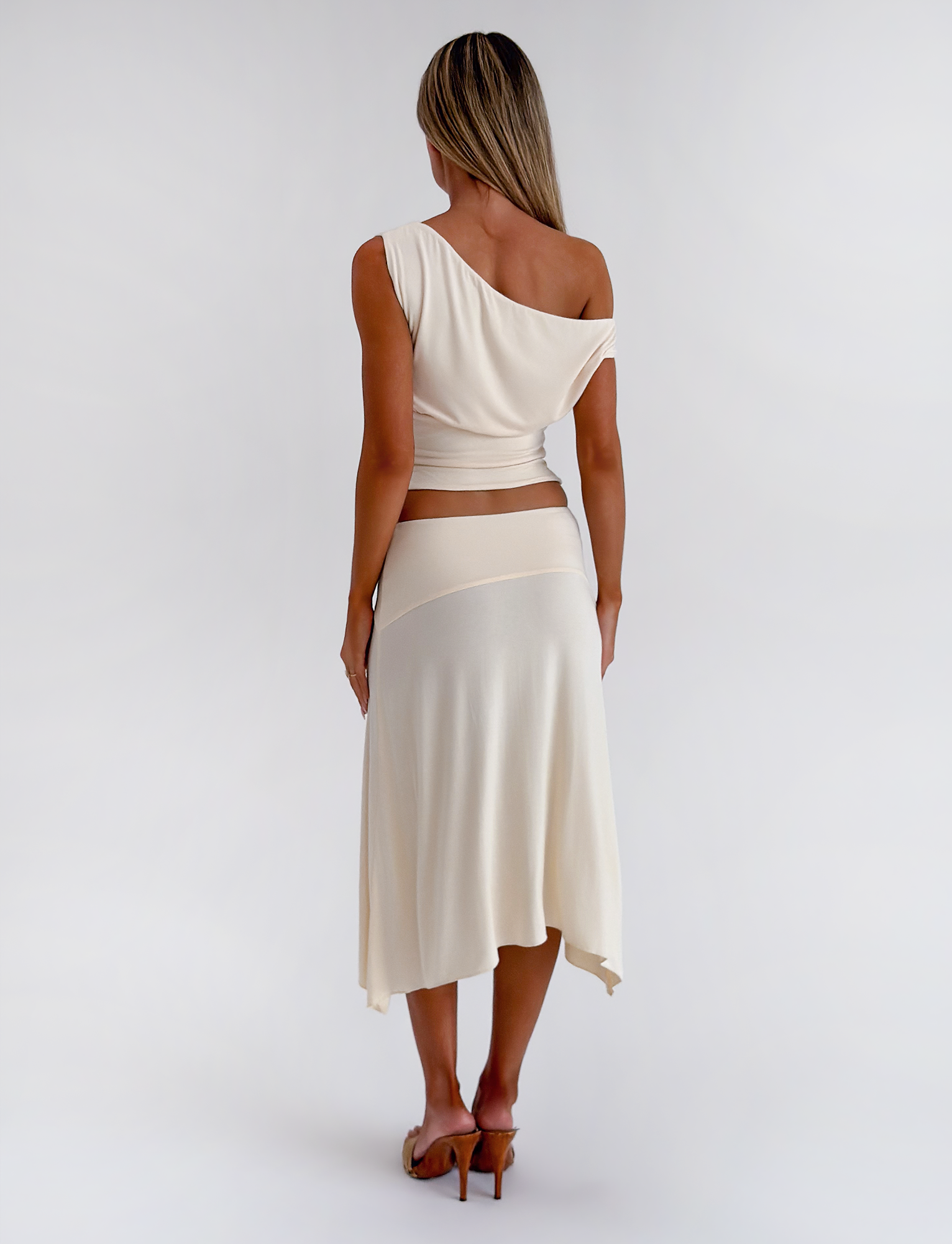 A woman faces away, wearing the TIGERMIST ALYSSA HI LO SKIRT in cream with a matching one-shoulder top and heeled sandals, set against a plain white background.