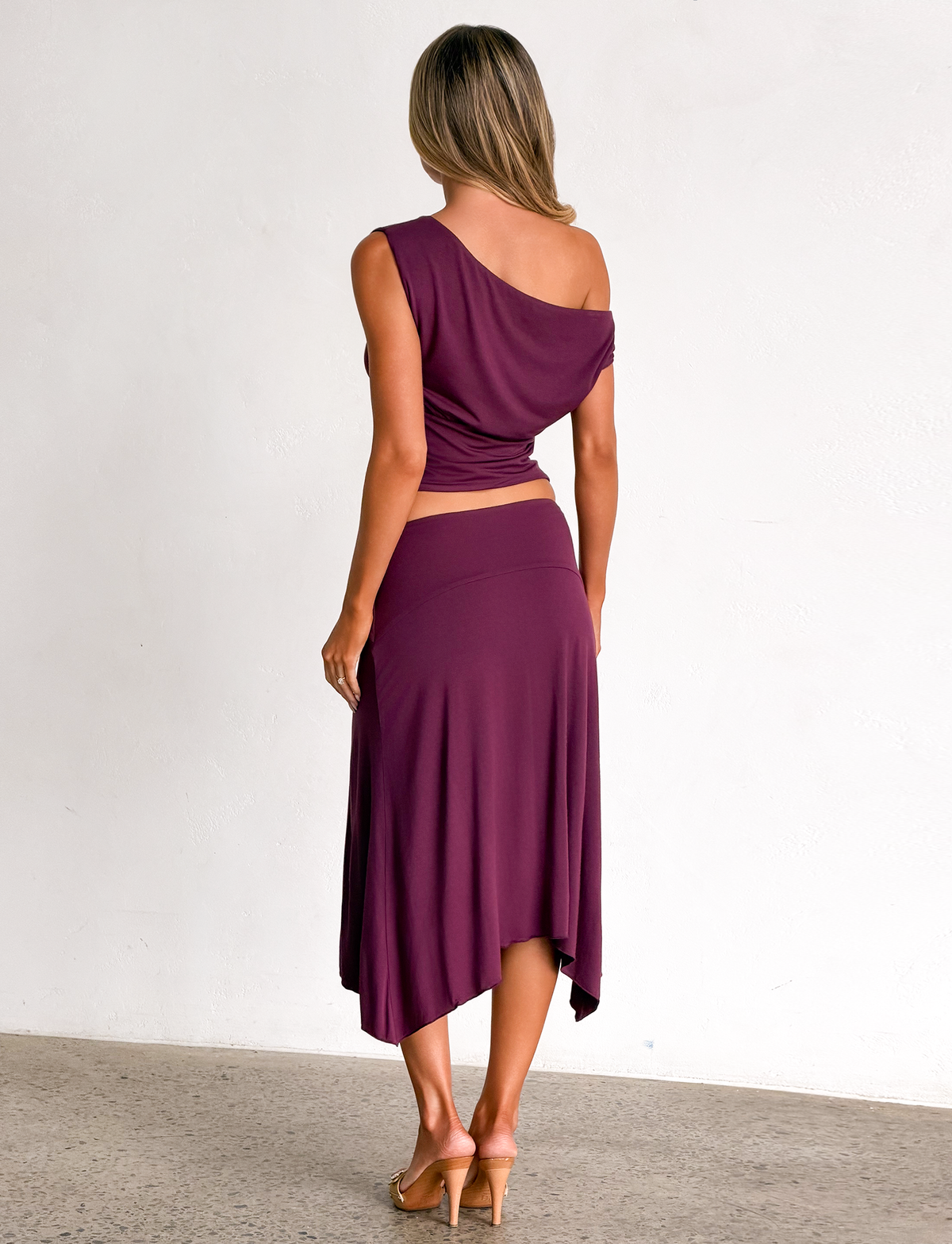 A woman stands facing away, wearing the TIGERMIST ALYSSA HI LO SKIRT in plum with tan heeled sandals, set against a plain light wall and concrete floor.