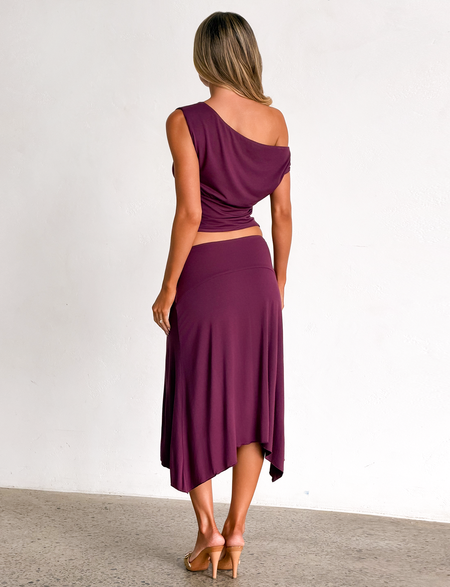 A woman stands facing away, wearing the TIGERMIST ALYSSA HI LO SKIRT in plum with tan heeled sandals, set against a plain light wall and concrete floor.