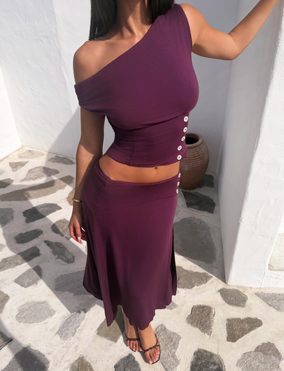 A woman stands outdoors on a stone patio near a white wall, modeling the TIGERMIST ALYSSA OFF SHOULDER TOP in plum—a matching set with a one-shoulder design, corseted waist, and skirt with side buttons. Her face is not visible.