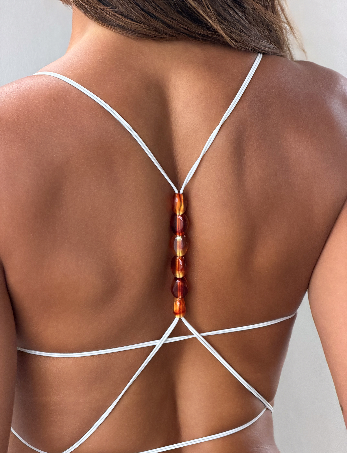 A close-up of a woman's back wearing the TIGERMIST MINERVA 2.0 BODYSUIT in cream lace, featuring strappy details and a vertical row of amber beads down the center.