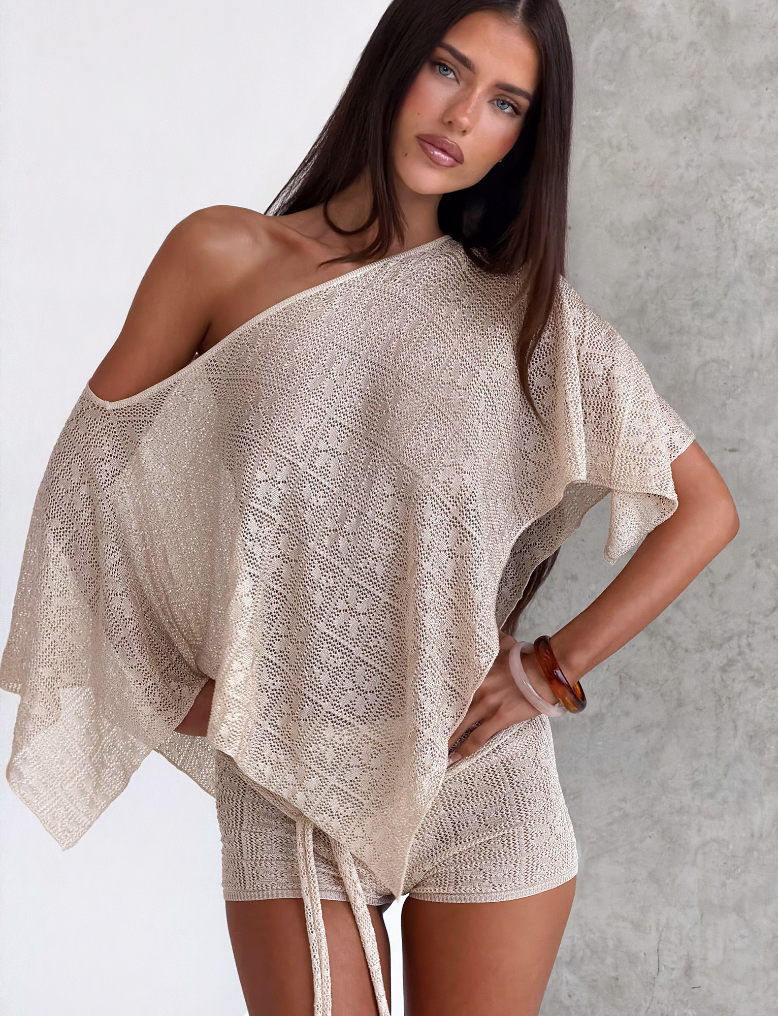 A woman with long dark hair wears the TIGERMIST FERN CROCHET PONCHO in champagne as a chic festival look, posing with one hand on her hip and a neutral expression against a light gray wall.